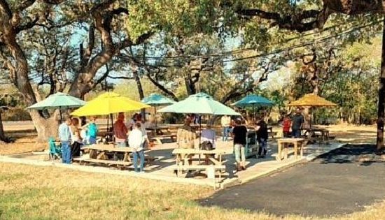 A Picture Of St. Nick's Outdoor Worship Service, Out On The Patio With Wooden Picnic Tables And Colorful Umbrellas Put Up, Between Trees That Also Shade The Patio.