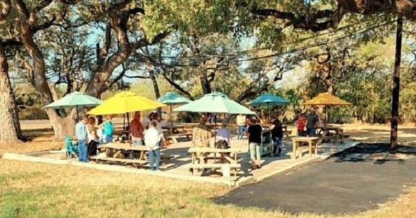 A Picture Of St. Nick's Outdoor Worship Service, Out On The Patio With Wooden Picnic Tables And Colorful Umbrellas Put Up, Between Trees That Also Shade The Patio.