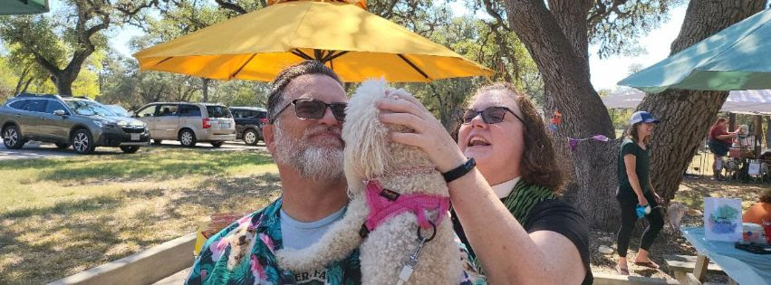 Woman holding up white fluffy dog and she and a man, both in sunglasses, are looking at the dog affectionately. Outside