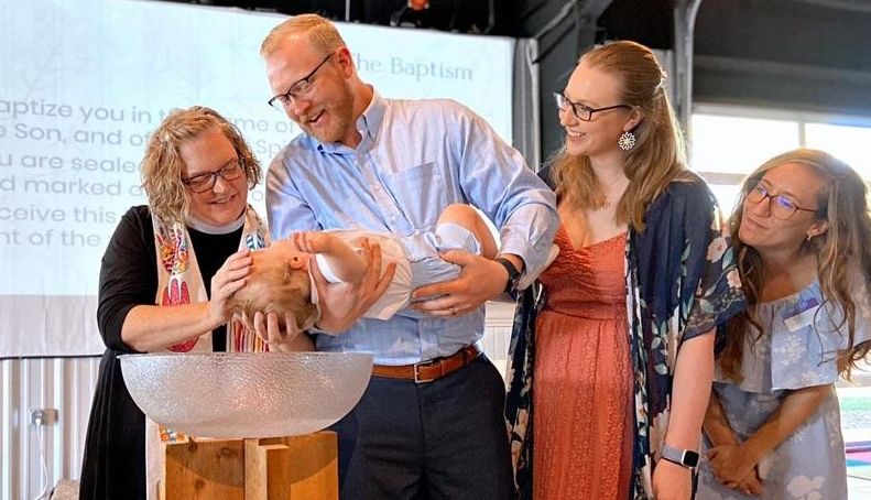 A woman in a black clergy shirt smiling and baptizing a baby held by a man with two other women looking on and smiling.