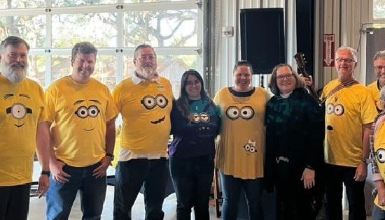 Seven Adults In Yellow Minion Shirts With Two Other Adults In Black, Lined Up Facing The Camera And Smiling
