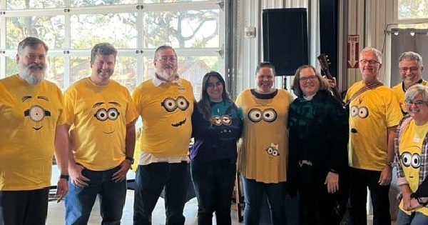 Seven Adults In Yellow Minion Shirts With Two Other Adults In Black, Lined Up Facing The Camera And Smiling