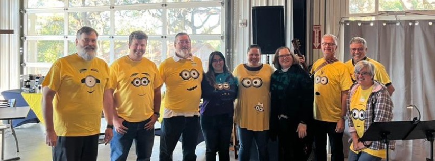 seven adults in yellow Minion shirts with two other adults in black, lined up facing the camera and smiling