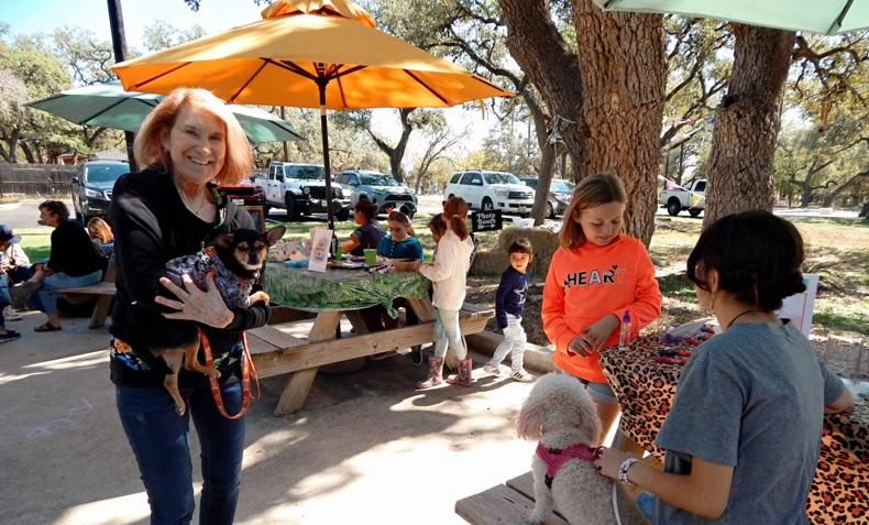 Older woman holding a dog and smiling at the camera with other people and pets around, outside, with picnic tables, on a sunny day.