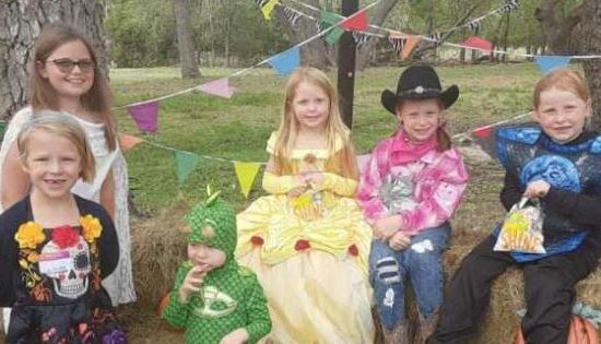 Children In Halloween Costumes And Smiling - Outside During The Day Between Some Trees With Some Hay Bales And Halloween Decorations