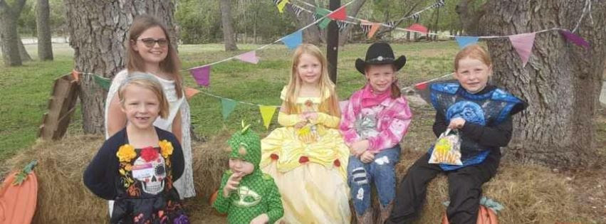 Children in Halloween costumes and smiling - outside during the day between some trees with some hay bales and Halloween decorations