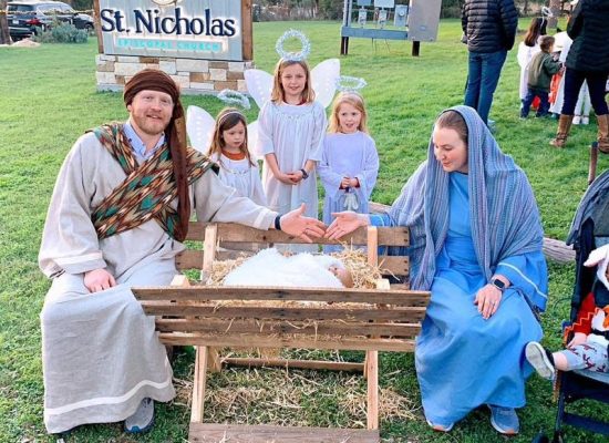 Two Adults And Three Children Dressed As Joseph, Mary And Angels, Sitting And Standing Around A Creche In An Outdoor Live Nativity Scene.