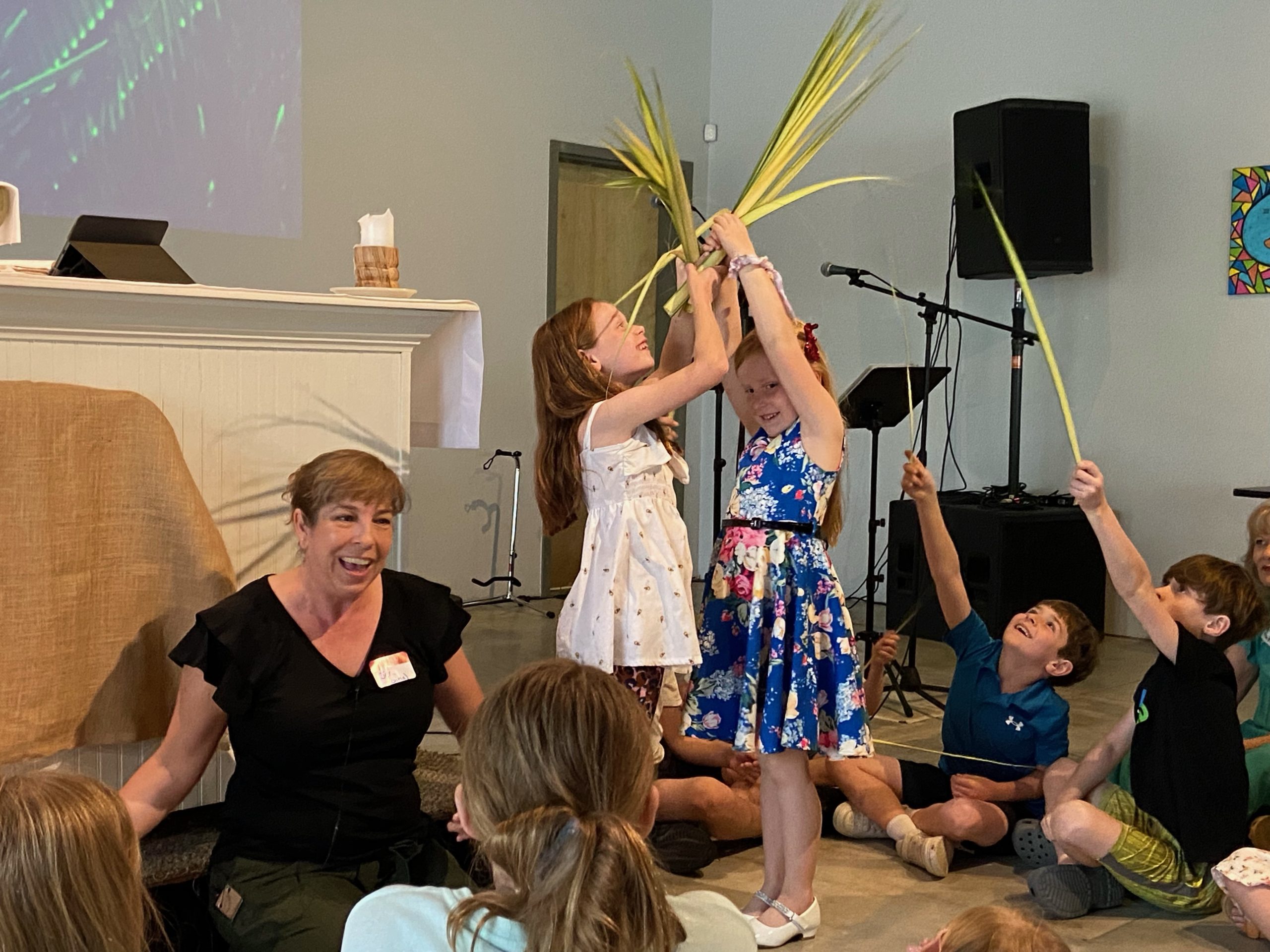 children waving palm leaves in the air and smiling with other children sitting on the floor in front of a smiling woman